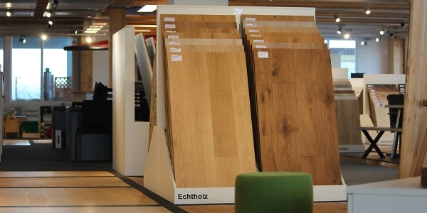 Ein Ausstellungsraum mit verschiedenen Echtholz-Bodenmustern, die auf Ständern präsentiert werden. Der Raum ist hell und modern gestaltet, mit Holzverkleidungen an den Wänden und einer Decke mit Spotlights. Ein grüner Hocker steht im Vordergrund.