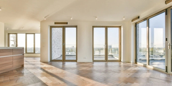 Ein lichtdurchfluteter Raum mit gro&szlig;en Fenstern und T&uuml;ren, die einen Panoramablick bieten. Der Boden ist mit elegantem Parkett ausgelegt, und die W&auml;nde sind in einem neutralen Wei&szlig; gehalten. Die Decke ist mit modernen Spotlights ausgestattet.