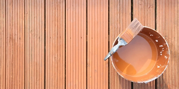 Ein Eimer mit brauner Farbe und ein Pinsel liegen auf einem Holzdeck. Die Farbe passt perfekt zum warmen Ton des Holzes, das in parallelen Linien verlegt ist. Ideal für Renovierungs- oder Verschönerungsprojekte im Außenbereich.
