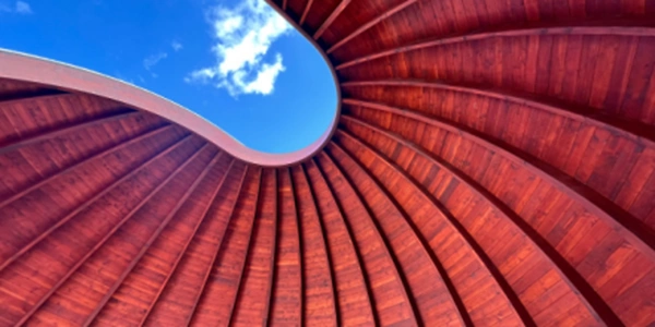 Eine geschwungene Holzstruktur mit rotbraunen Paneelen, die einen eleganten Bogen bilden. Durch die Öffnung ist der blaue Himmel sichtbar. Die Konstruktion zeigt ein harmonisches Zusammenspiel von Architektur und Natur.