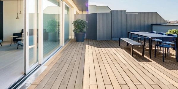 Eine gro&szlig;z&uuml;gige Terrasse mit Holzboden, die an einen Innenraum mit gro&szlig;en Glas-Schiebet&uuml;ren anschlie&szlig;t. Die Terrasse ist mit einem Tisch und St&uuml;hlen ausgestattet und bietet viel Platz f&uuml;r Entspannung. Ein Pflanzk&uuml;bel sorgt f&uuml;r einen gr&uuml;nen Akzent.